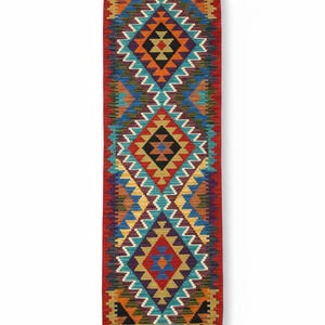 May include: A rectangular woven runner rug featuring a vibrant geometric pattern with repeating diamond shapes in red, orange, yellow, blue, purple, and teal. The rug has a serrated zigzag border and short, light-colored fringe tassels at both ends.