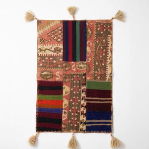 May include: A rectangular wall hanging rug featuring a patchwork design of various woven textile segments in shades of red, green, blue, black, and beige. The rug is finished with six beige tassels attached to the corners and the center edges.