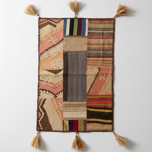 Peut inclure: Une tapisserie murale rectangulaire composée d'un assemblage de divers motifs textiles tissés dans des tons de rose, marron, gris, rouge et bleu. La pièce est finie avec des bords cousus foncés et six pompons beiges fixés aux coins et sur les côtés.