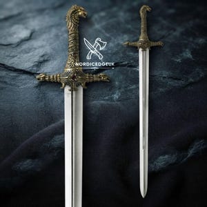 Può includere: Una spada in metallo con lama color argento e un'elsa decorata color bronzo, caratterizzata da un pomolo a forma di testa di lupo. Il testo NORDICEDGEUK è visibile al centro. Il design presenta dettagli intricati e una piccola pietra rossa.
