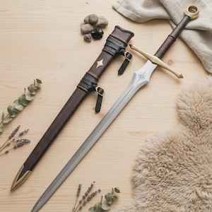 Peut inclure: Une épée en métal argenté avec une garde et un pommeau dorés, présentant une gravure en forme d'étoile sur la lame. Elle est accompagnée d'un fourreau en cuir marron foncé avec des lanières noires, des boucles dorées et un emblème étoilé.