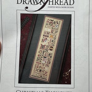 May include: A printed pattern sheet for The Drawn Thread Christmas Thoughts Sampler, featuring a vertical alphabet design with holiday motifs in red, green, and black on a cream background, framed in a dark border with small gold scissors.