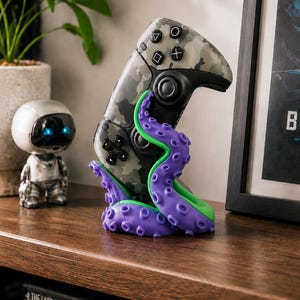 Può includere: Un controller per videogiochi con motivo mimetico tenuto in posizione verticale da un supporto a forma di tentacolo di polpo viola e verde. Il controller presenta pulsanti e joystick neri, mentre la base del tentacolo mostra ventose strutturate.