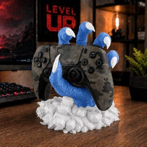Può includere: Un controller per videogiochi mimetico nero e grigio tenuto da una mano di mostro blu testurizzata con artigli bianchi affilati. La mano emerge da una base di pezzi bianchi, frastagliati e simili a rocce.