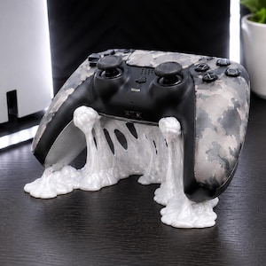 May include: A grey and black camouflage patterned video game controller resting on a white, glossy, melted-look stand. The controller features two black analogue sticks, a directional pad, and four action buttons on the right side of the device.