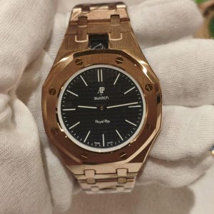 Puede incluir: Un reloj de pulsera en tono oro rosa con un bisel octogonal que presenta ocho tornillos visibles. La esfera negra tiene un patrón texturizado, marcadores de hora blancos y el texto AP, swatch y Royal Rop impreso en blanco. Correa metálica.