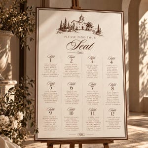 May include: A rectangular wedding seating chart on a wooden easel with a cream background and dark brown text. The top features a sketch of a villa and the text "PLEASE FIND YOUR SEAT". It lists twelve tables with guest names in a grid layout.