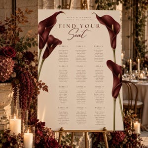 Puede incluir: Un cartel de distribución de asientos de boda sobre un caballete con el texto "Maya & George" y "Find Your Seat" junto a una lista de doce mesas y nombres de invitados. El cartel está decorado con ilustraciones de calas color burdeos.
