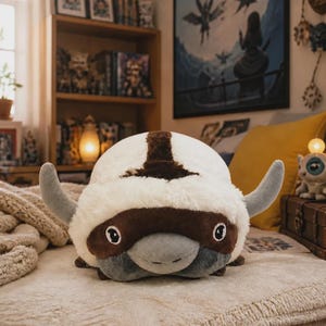 Pode incluir: Um peluche em forma de bisão com pelo cor de creme, uma marca castanha escura em forma de seta na cabeça, um focinho cinzento e dois chifres cinzentos curvos. A criatura tem grandes olhos pretos e brancos e um corpo arredondado.