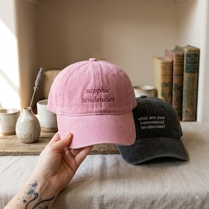 Op de afbeelding: Een oudroze baseballpet met de geborduurde tekst "sapphic tendencies" in een donker lettertype, geplaatst voor een antracietgrijze baseballpet met de geborduurde tekst "what are your homosexual tendencies?" in een licht lettertype.