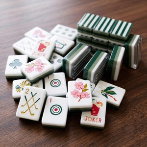 May include: A collection of white mahjong tiles featuring various colourful illustrations including pink cherry blossoms, red cocktails labelled JOKER, green golf clubs, and bullseye targets, alongside several tiles stacked with green striped backs.