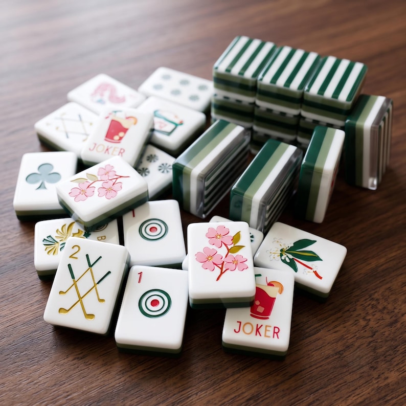 May include: A collection of white mahjong tiles featuring various colourful illustrations including pink cherry blossoms, red cocktails labelled JOKER, green golf clubs, and bullseye targets, alongside several tiles stacked with green striped backs.