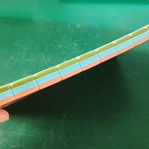 May include: A rectangular object featuring a layered design with three distinct horizontal bands. The top layer is olive green, the middle layer is light blue, and the bottom layer is a soft peach color. The surface has a grid of small square segments.