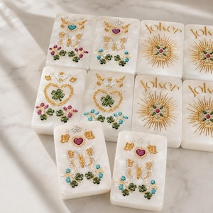May include: A set of rectangular white tiles with a pearlescent finish. Some tiles feature gold-tone butterflies, hearts, and clover motifs with colorful accents. Other tiles display the word Joker in gold script above a sunburst design.