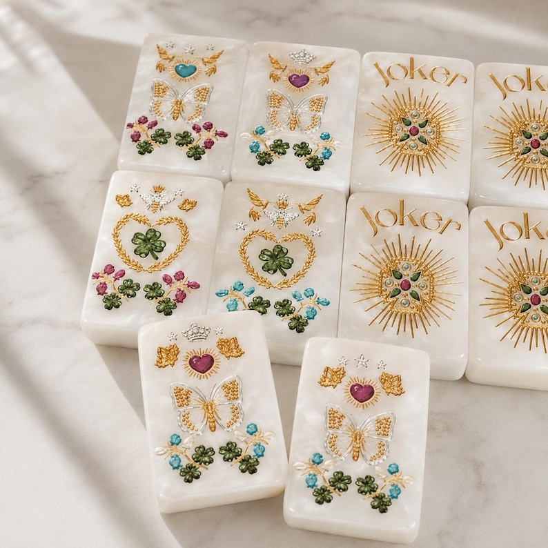 May include: A set of rectangular white tiles with a pearlescent finish. Some tiles feature gold-tone butterflies, hearts, and clover motifs with colorful accents. Other tiles display the word Joker in gold script above a sunburst design.