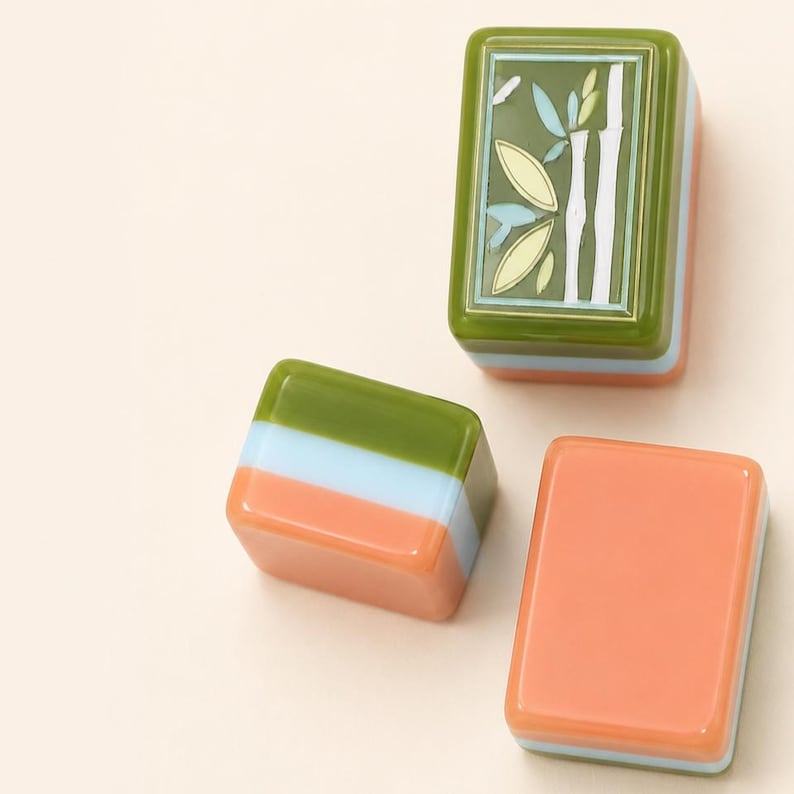 May include: Three rectangular mahjong tiles with smooth, glossy surfaces. One tile features a green bamboo design on a dark green background. The other two tiles show horizontal layers of olive green, light blue, and peach orange colors.