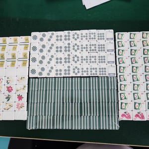 May include: A set of white mahjong tiles arranged in groups. The tiles feature various designs including the word NEWS, floral patterns, joker icons, numbered suits with circular and star motifs, and green striped backs. Some tiles display red dragon symbols.