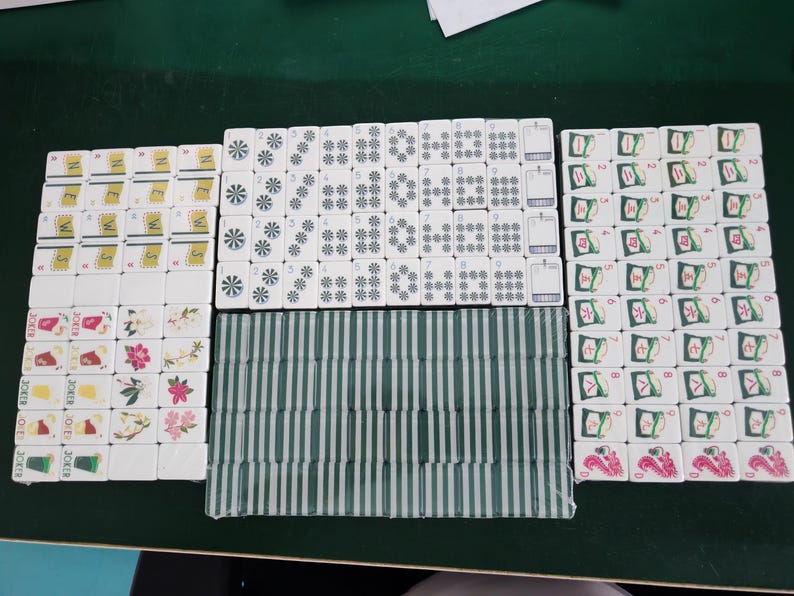 May include: A set of white mahjong tiles arranged in groups. The tiles feature various designs including the word NEWS, floral patterns, joker icons, numbered suits with circular and star motifs, and green striped backs. Some tiles display red dragon symbols.