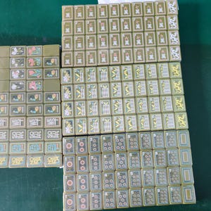May include: A set of olive green mahjong tiles arranged in rectangular blocks. The tiles feature various engraved designs including Chinese characters, bamboo stalks, circular patterns, floral motifs, and directional labels like WEST and SOUTH.