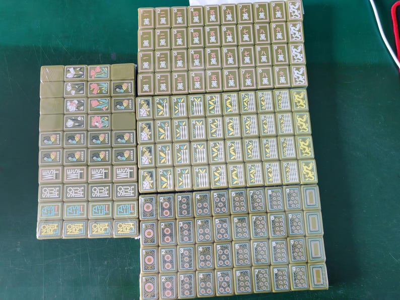 May include: A set of olive green mahjong tiles arranged in rectangular blocks. The tiles feature various engraved designs including Chinese characters, bamboo stalks, circular patterns, floral motifs, and directional labels like WEST and SOUTH.