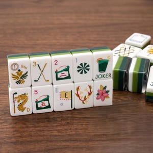 May include: A set of white and green mahjong tiles featuring colourful illustrations including a dragon, golf clubs, a cocktail glass labelled JOKER, a pink flower, and various numbers and letters. The tiles are arranged in a rectangular formation.