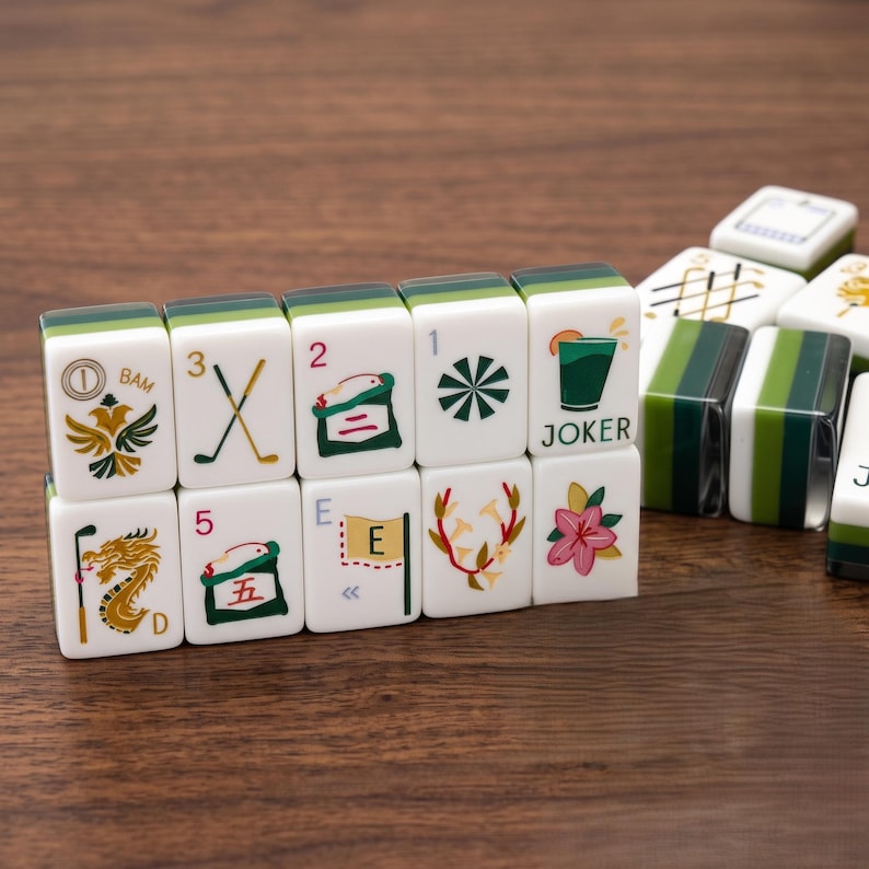 May include: A set of white and green mahjong tiles featuring colourful illustrations including a dragon, golf clubs, a cocktail glass labelled JOKER, a pink flower, and various numbers and letters. The tiles are arranged in a rectangular formation.