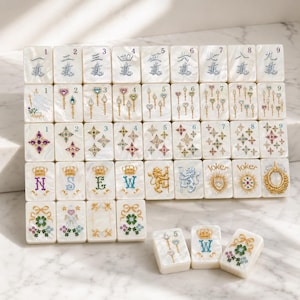 May include: A set of rectangular mahjong tiles with a pearlescent white finish. The tiles feature intricate, colorful illustrations of scepters, stars, crowns, letters N, S, E, W, lions, and floral motifs. Some tiles display numbers 1 through 9.