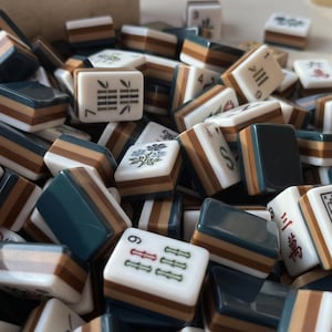 May include: A collection of rectangular mahjong tiles featuring a layered design with a white top, a brown middle stripe, and a dark teal base. The tiles display various engraved symbols, including bamboo patterns, floral motifs, and Chinese characters.