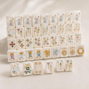 May include: A set of white pearlescent mahjong tiles featuring intricate engraved designs, including Chinese numerals, colorful star patterns, floral motifs, crowns, lions, and joker icons with gold, silver, and multi-colored accents throughout.