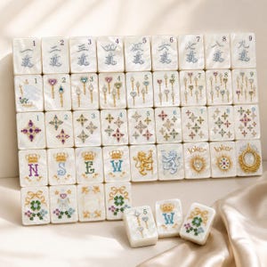 May include: A set of rectangular mahjong tiles with a pearlescent white finish. The tiles feature intricate, colorful illustrations including numbers, ornate scepters, star-shaped gems, crowns, letters N, S, E, W, lions, and floral motifs with bows.