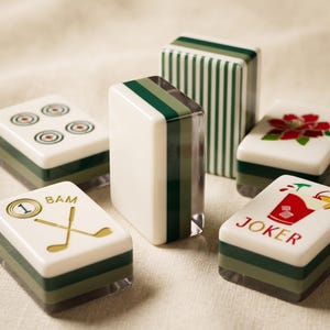 May include: A collection of rectangular mahjong tiles featuring white faces with various designs including a red cocktail graphic labelled JOKER, a gold golf club icon with the text 1 BAM, a red flower, and circular patterns, with green striped sides.