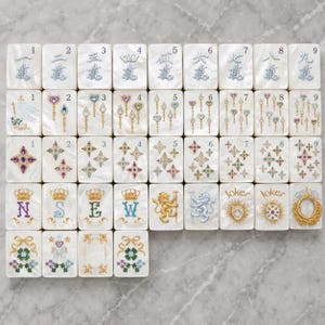 May include: A set of white rectangular mahjong tiles arranged in a grid. The tiles feature colorful illustrations of numbers, wands, stars, crowns with letters N, S, E, W, lions, joker text, and floral motifs with ribbons in various metallic and jewel tones.