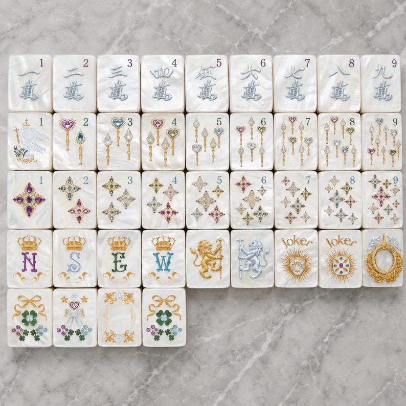 May include: A set of white rectangular mahjong tiles arranged in a grid. The tiles feature colorful illustrations of numbers, wands, stars, crowns with letters N, S, E, W, lions, joker text, and floral motifs with ribbons in various metallic and jewel tones.