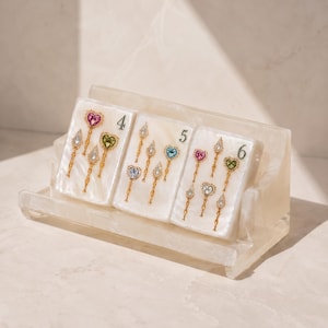 May include: A white acrylic display stand holding three rectangular white tiles, each labeled with the numbers 4, 5, and 6. Each tile features small gold-toned pins with colorful heart and teardrop-shaped gemstones in pink, blue, green, and clear.
