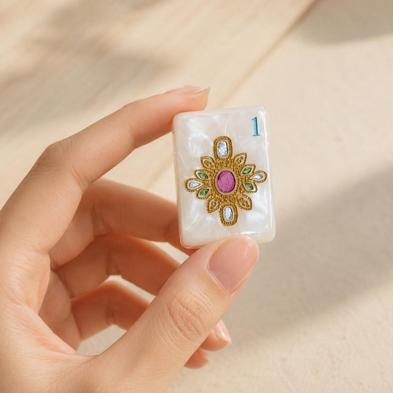 May include: A rectangular white pearlescent mahjong tile featuring a central gold filigree emblem with a pink oval center, surrounded by small blue and green accents, and the number 1 printed in blue in the upper right corner of the tile.