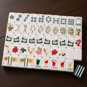May include: A set of white mahjong tiles arranged in a grid, featuring various colourful illustrations including numbers, golf clubs, flowers, dragons, the letters N, E, W, S on flags, and glasses labelled JOKER, with one tile showing green stripes.