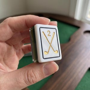 May include: A rectangular white game tile featuring a black border and a gold graphic of two crossed golf clubs with the number 2 positioned above them. The tile has a smooth, glossy finish and a dark green base, held between fingers.