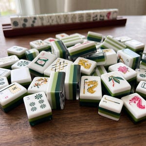 May include: A collection of white and green mahjong tiles featuring various engraved symbols, including dragons, flowers, and numbers. Some tiles are arranged in a wooden rack in the background, while others are scattered in the foreground.