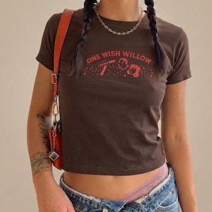 May include: A dark brown short-sleeve t-shirt featuring the red text ONE WISH WILLOW printed across the chest, accompanied by small red graphic illustrations of a wand, a star, and a small character, paired with blue denim jeans and a red bag.