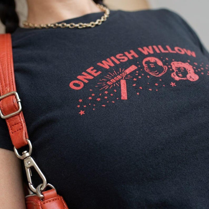May include: A black t-shirt featuring red graphic text that reads ONE WISH WILLOW, accompanied by illustrations of a firecracker, stars, and two cartoon faces. The shirt is worn with a gold chain necklace and an orange bag with a metal clasp.
