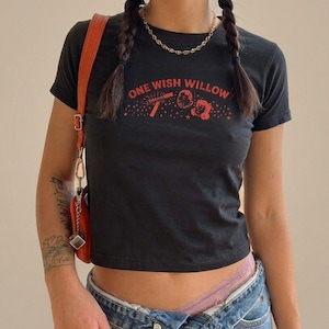 May include: A black short-sleeved t-shirt featuring the red text ONE WISH WILLOW printed across the chest, accompanied by a graphic illustration of a magic wand and two cartoon faces surrounded by small stars.