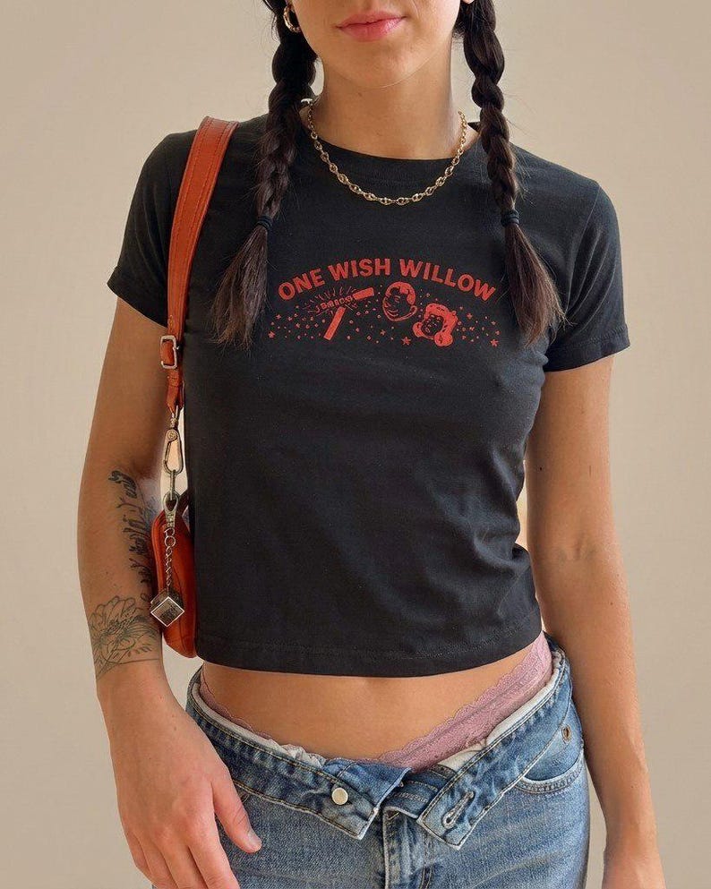 May include: A black short-sleeved t-shirt featuring the red text ONE WISH WILLOW printed across the chest, accompanied by a graphic illustration of a magic wand and two cartoon faces surrounded by small stars.