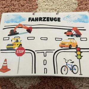 May include: A laminated educational card titled FAHRZEUGE featuring illustrations of a construction excavator, a dump truck, a road roller, a yellow car, a bicycle, a red stop sign, a traffic light, and an orange traffic cone on a road layout.