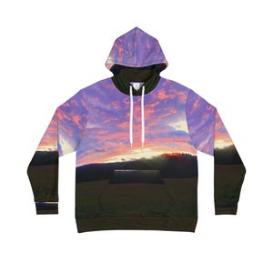 Puede incluir: Una sudadera con capucha con un diseño de puesta de sol. La sudadera muestra un cielo con tonos morados, rosas y naranjas, y un paisaje verde oscuro en la parte inferior. Tiene una capucha interior negra y cordones blancos.