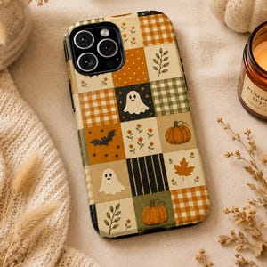 May include: A smartphone case featuring a patchwork design of autumn-themed squares. The pattern includes white ghosts, orange pumpkins, black bats, maple leaves, floral sprigs, and orange or green gingham and striped squares on a cream background.
