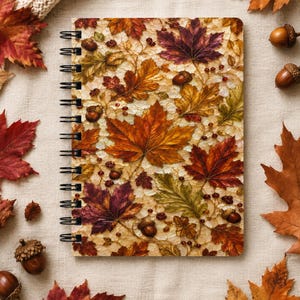Pode incluir: Um caderno de espiral com um padrão detalhado na capa com tema de outono, apresentando folhas de bordo em tons de laranja, vermelho, amarelo e castanho, intercaladas com pequenas bolotas castanhas e bagas sobre um fundo claro e texturado.