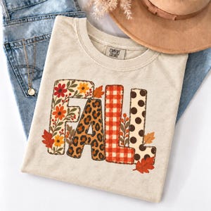 May include: A beige short-sleeve t-shirt with the word FALL printed in large letters. The F features a floral pattern, the A has a leopard print, the L is red and white gingham, and the second L has brown polka dots with autumn leaves.