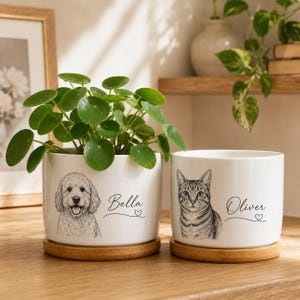 Peut inclure: Deux pots de fleurs cylindriques en céramique blanche avec des soucoupes en bois. Le pot de gauche présente un dessin au trait d'un chien et le nom Bella. Le pot de droite présente un dessin au trait d'un chat et le nom Oliver. Une plante verte est dedans.