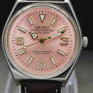 May include: A silver-tone wristwatch with a pink dial, featuring the text SEIKO, OYSTER PERPETUAL, EXPLORER, and SUPERLATIVE CHRONOMETER OFFICIALLY CERTIFIED. The watch has a brown leather strap, silver hands, and white-filled hour markers.