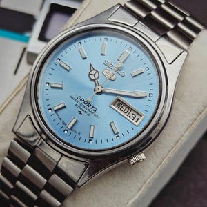 May include: A Seiko 5 Sports automatic watch with a light blue dial, silver-tone hands, and hour markers. The watch features a day-date window at the three o'clock position, a stainless steel case, and a matching link bracelet. Text on the dial reads Seiko 5 Sports, Water 10 Bar Resist, Automatic, and Japan 7009-876L R.